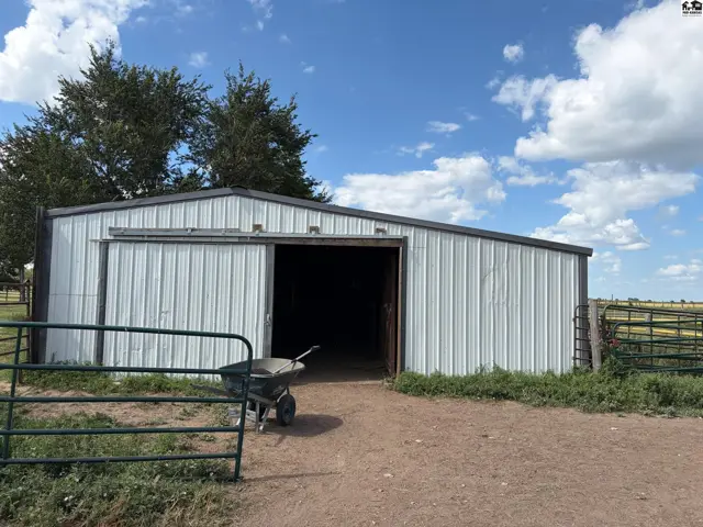 Metal barn with sliding door, featuring a wheelbarrow and rural landscape under a cloudy sky.
