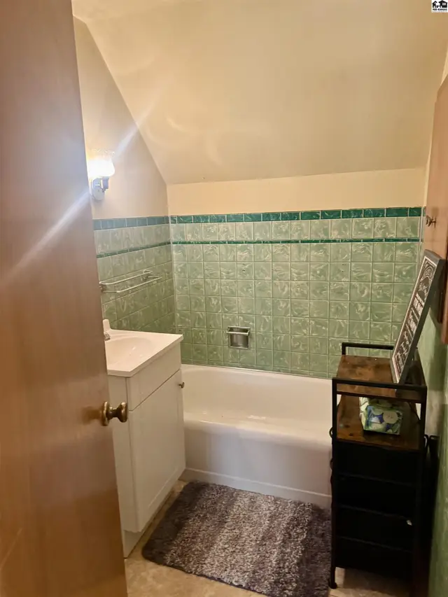 Compact bathroom features a white tub, vanity, and green tiled walls under a sloped ceiling.