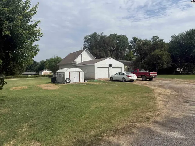 White house with attached garage, storage shed, and vehicles on a grassy lot.