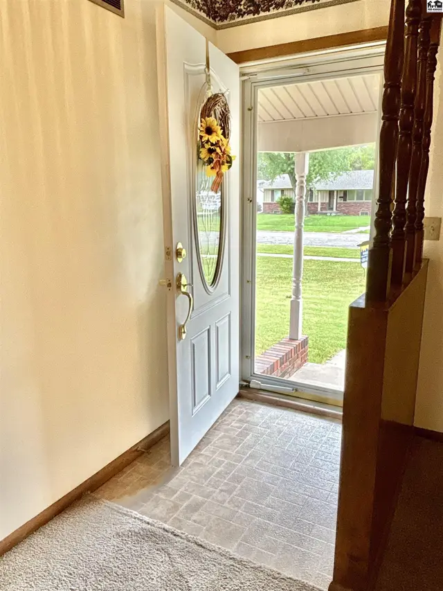 Open white front door with oval window, revealing a sunlit entryway with wood banister and view of a suburban street.