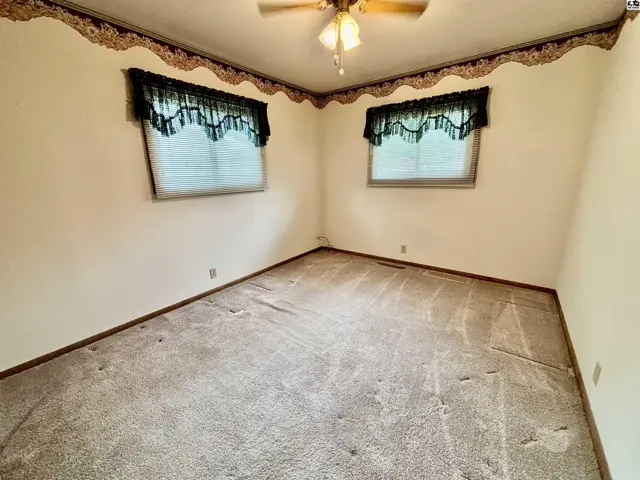 Empty bedroom with beige carpet, white walls, decorative wallpaper border, and two windows with blinds.