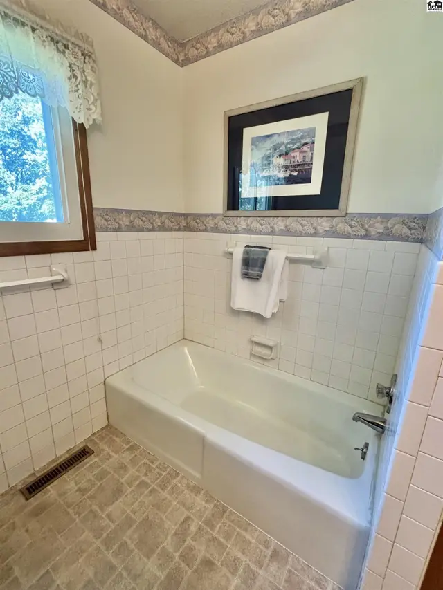 White tiled bathroom with a bathtub, patterned wallpaper, and a framed picture above the tub.