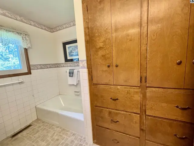 Bathroom with white tiled tub surround, patterned wallpaper, and a large, built-in wooden storage cabinet.
