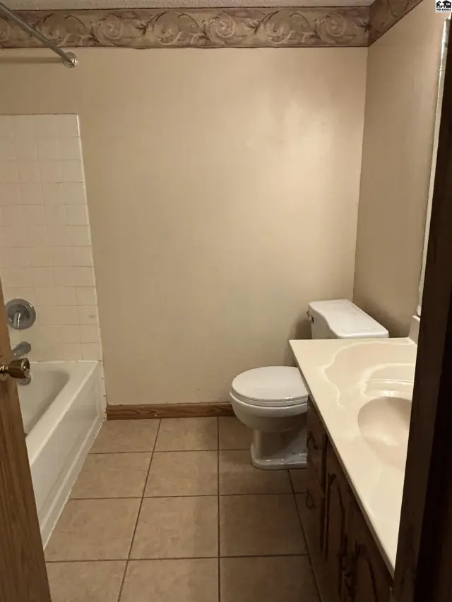 A neutral bathroom features a white tub, toilet, and vanity with tiled flooring and decorative wallpaper.
