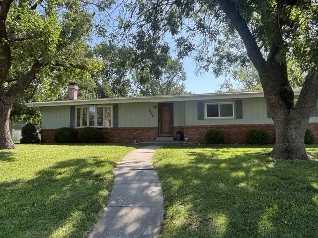 Exterior of a light green ranch home with brick accents, a bay window, and mature trees.