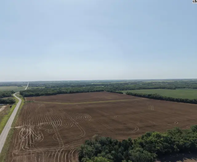 Expansive aerial view of tilled farmland with a paved road and distant tree line under a clear sky.