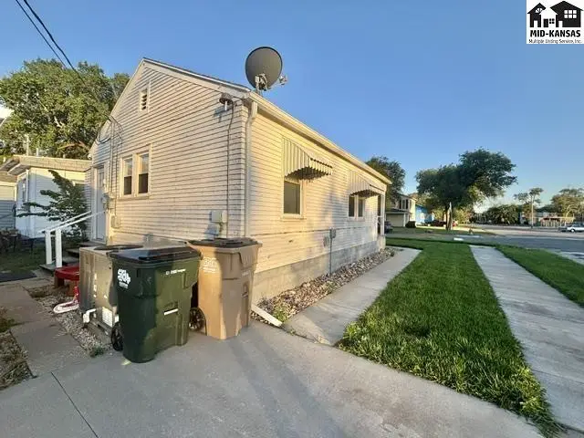 Exterior view of a light-colored vinyl-sided house with awnings, concrete driveway, and green lawn.