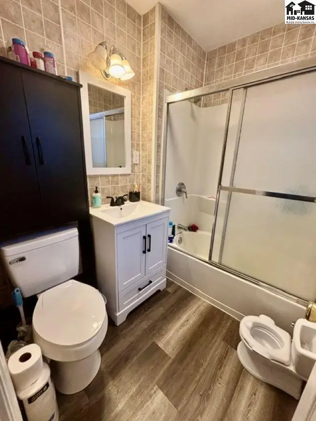 Compact bathroom with white vanity, tiled walls, wood-look flooring, and a sliding glass shower door.