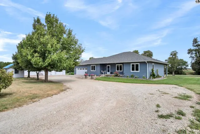 Blue ranch home with a gravel driveway, mature trees, and a detached garage on a sunny day.