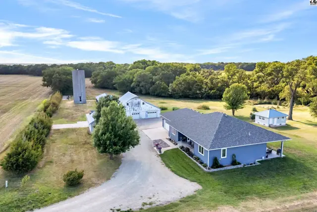 Sprawling rural property features a blue ranch home, white barn, and silo surrounded by trees.