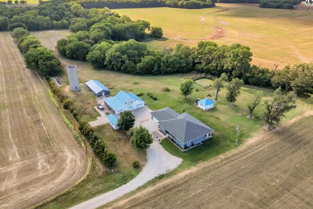 Aerial view of a rural property featuring a blue-roofed house, silo, barns, and outbuildings surrounded by fields.