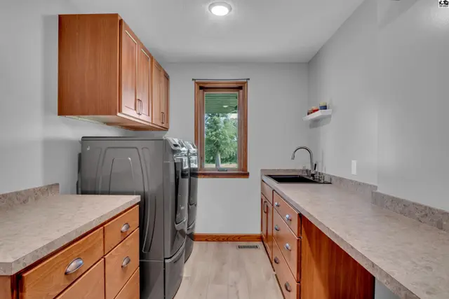 Spacious laundry room with gray appliances, wood cabinetry, granite countertops, and a deep sink.