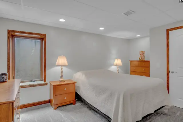 Cozy basement bedroom with a queen bed, wood-trimmed window, and light gray walls.