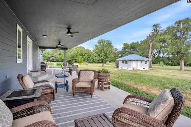 Spacious covered patio with wicker furniture and ceiling fans overlooks a large, tree-filled yard.