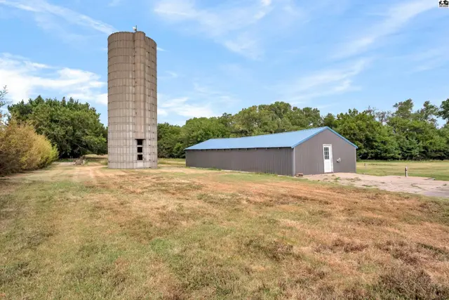 Concrete silo and gray metal building with blue roof stand on a grassy lot under a blue sky.