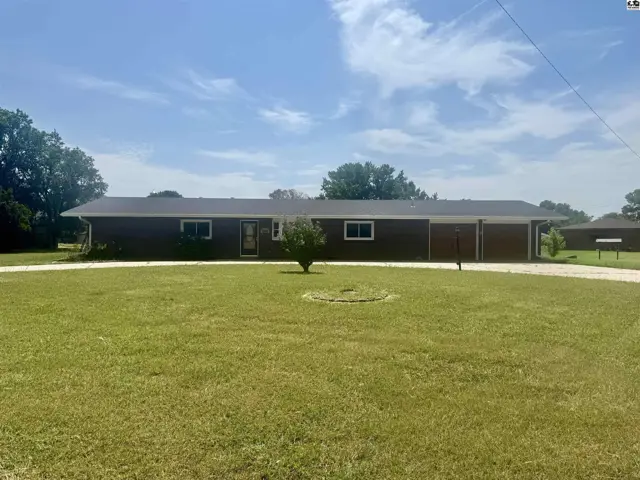 This single-story home features a dark brown exterior, a wide driveway, and a large, manicured lawn.