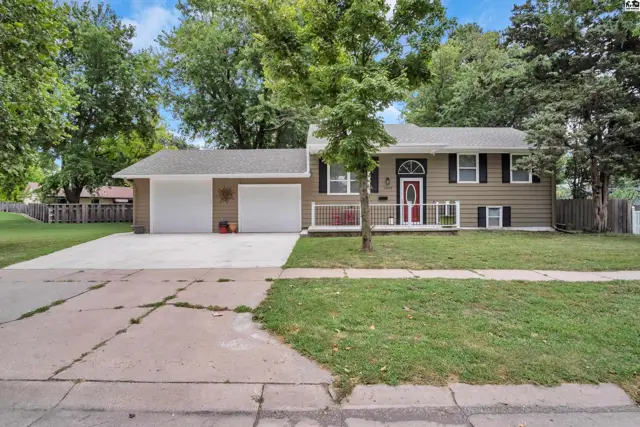 This tan-sided home features a red front door, two garage doors, and mature trees.