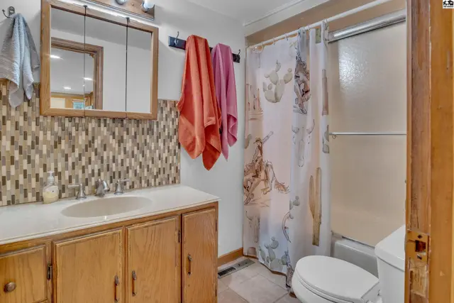 This bathroom features a wooden vanity, tiled backsplash, and a shower with a patterned curtain.