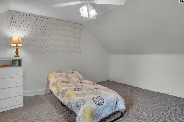 This attic bedroom features a ceiling fan, carpeted floor, and a dresser.