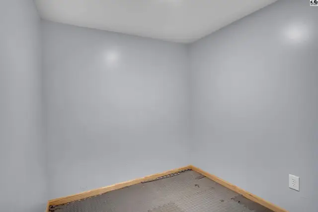 This empty room features light gray walls, a wood-trimmed floor, and a white electrical outlet.