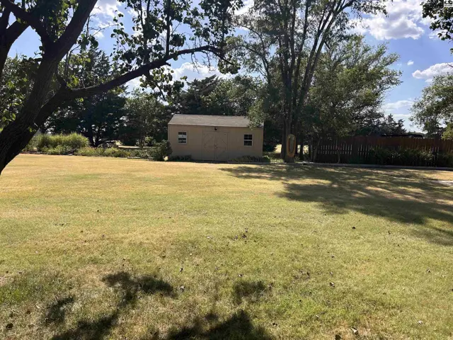 A tan shed with two windows sits in a large, grassy yard with mature trees.