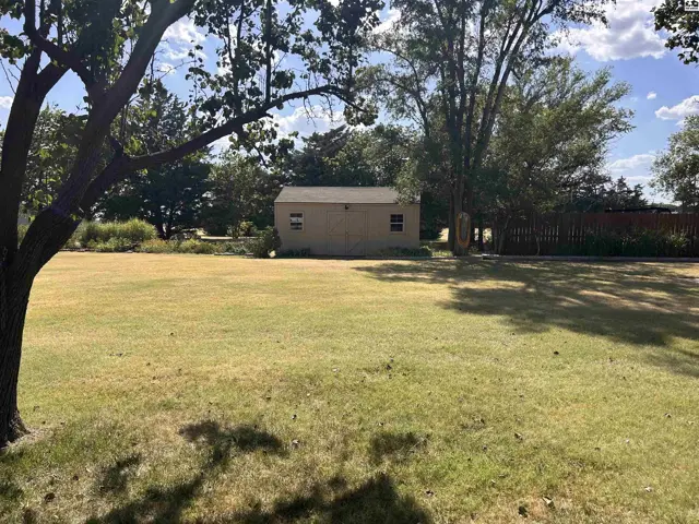 A tan shed with two windows sits in a large, grassy yard with mature trees.