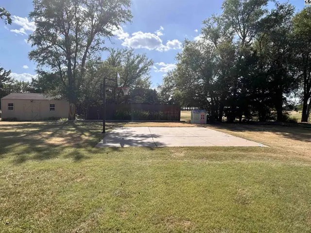 Concrete basketball court with a hoop is surrounded by a grassy lawn and mature trees.