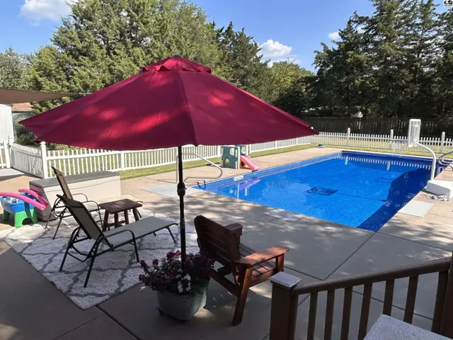Inground swimming pool with concrete deck, patio furniture, and white picket fence.