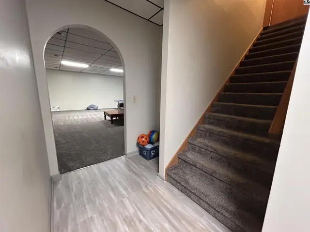 Carpeted stairs ascend beside a doorway opening into a finished room with wood-look flooring.