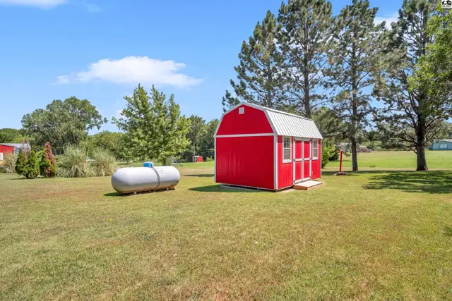 A red storage shed with a white metal roof sits on a grassy lawn next to a propane tank.