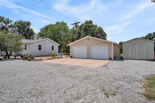 A white house, a two-car garage, and a storage shed sit on a gravel lot.