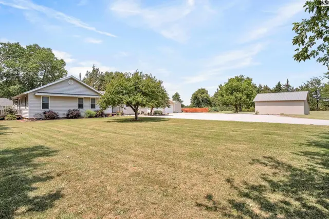 A light-colored single-story home sits on a large, grassy lot with mature trees and a detached garage.