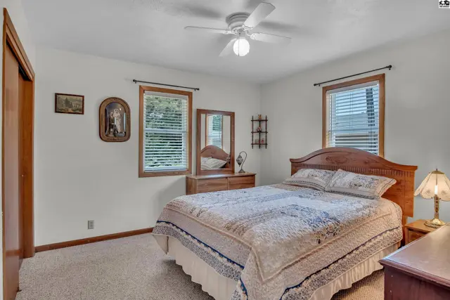 This bedroom features a ceiling fan, carpeted floor, and two windows with blinds.