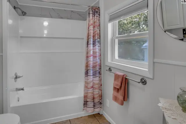 White bathtub with shower surround, patterned shower curtain, and a window with a towel rack.