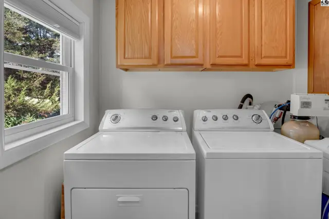 White washer and dryer units sit beneath light wood cabinets in a room with a large window.