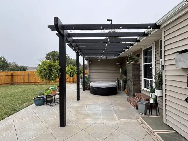 A black wooden pergola with string lights covers a concrete patio with a hot tub.