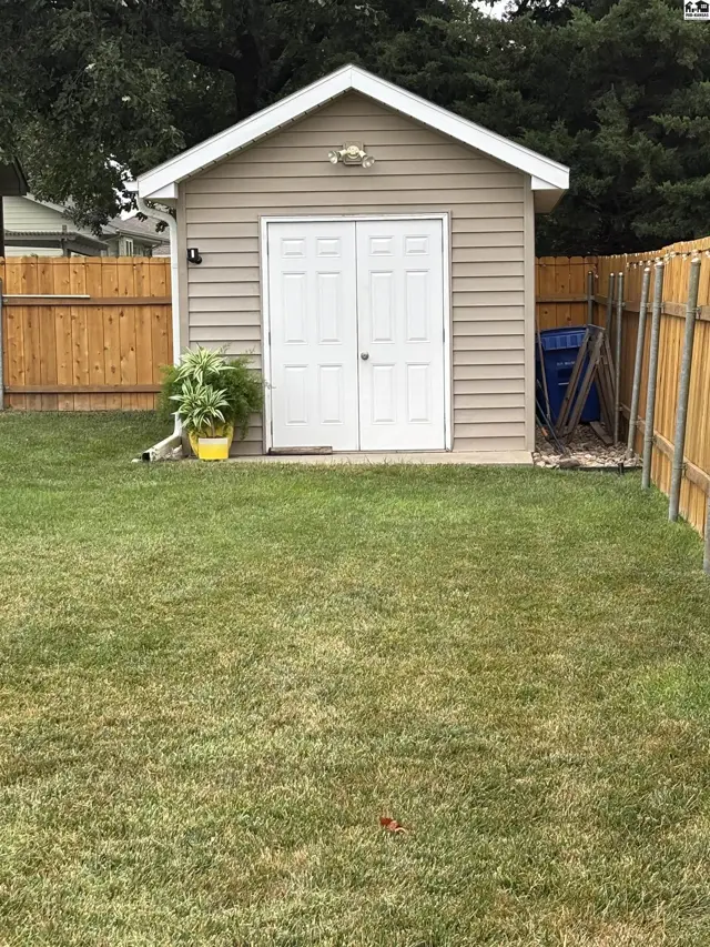 A tan vinyl-sided shed with double white doors sits on a green lawn.