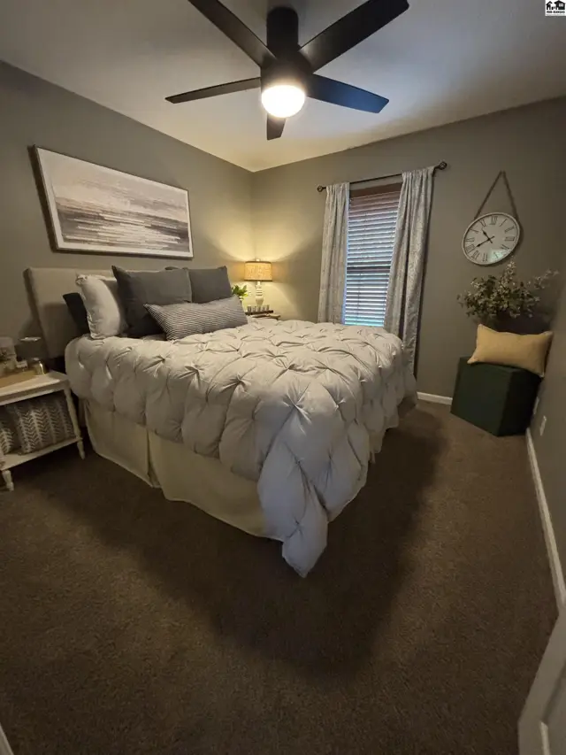 This bedroom features carpeted floors, a ceiling fan, and a large window with blinds.