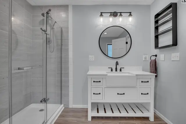 Modern bathroom features a glass-enclosed shower, white vanity with black hardware, and a round mirror.