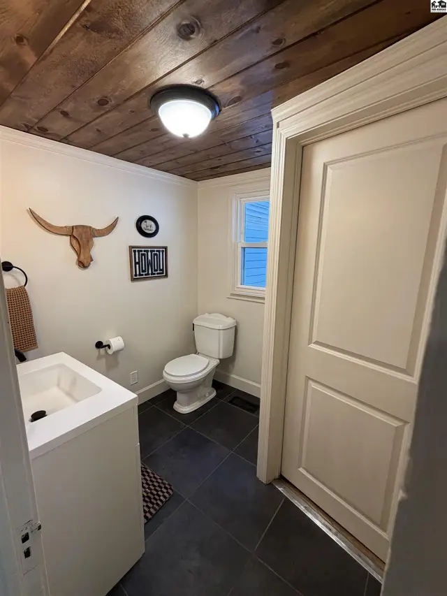 This bathroom features a white vanity, toilet, dark tile flooring, and a wood-plank ceiling.