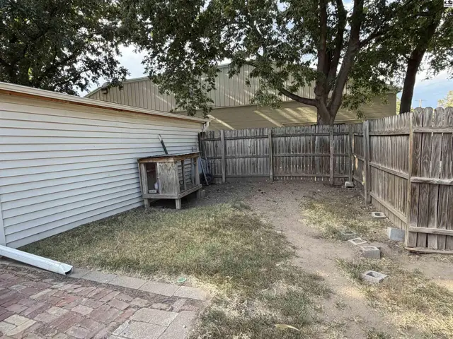 A wooden dog house sits in a fenced yard with a brick patio.