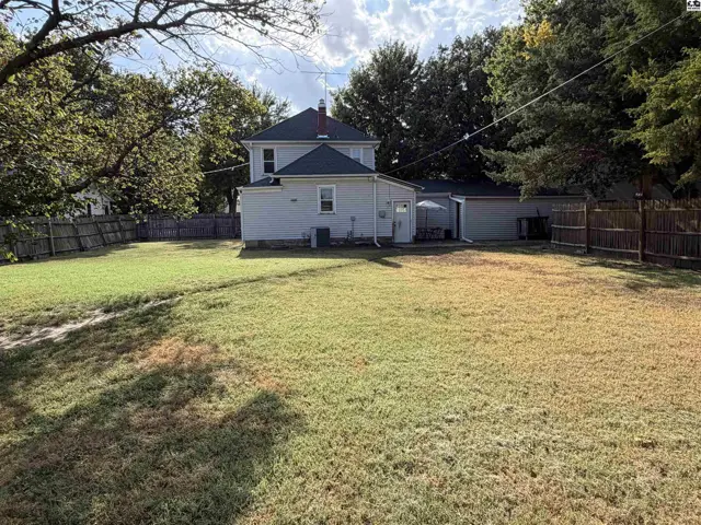 A light-colored house with a dark roof sits on a grassy lot with mature trees.