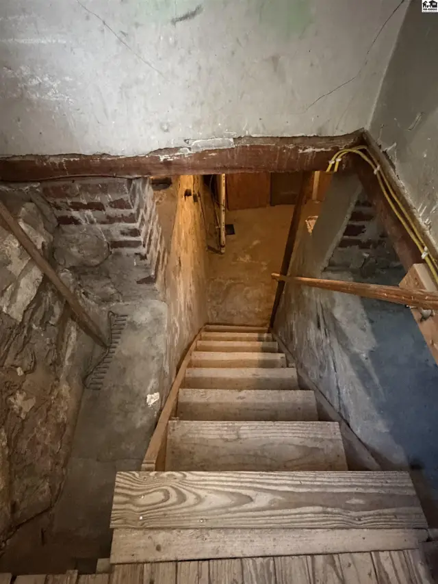 Wooden stairs descend into a basement with stone and concrete walls.