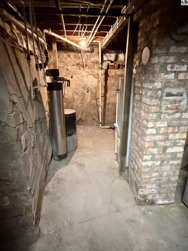 Concrete floor and brick walls in a basement with exposed pipes and a water softener.