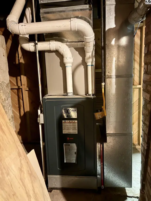 Furnace with metal ductwork and white PVC pipes against wood and brick walls.