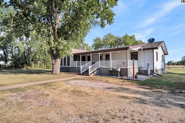 White siding ranch home with a covered wraparound porch and mature trees.