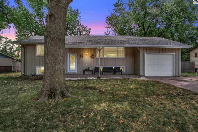 This tan-sided home features a covered porch, attached garage, and mature trees.