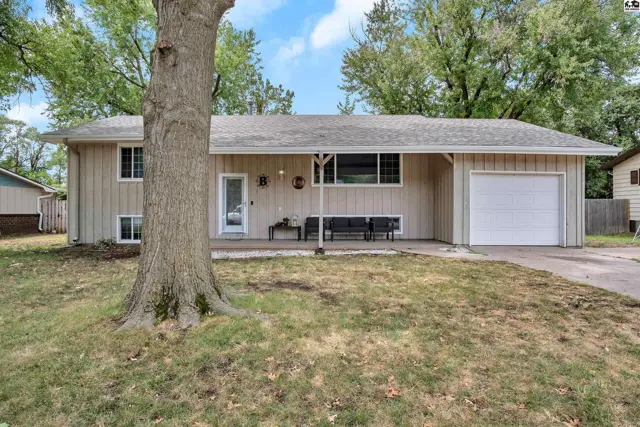 This tan-sided home features a covered porch, attached garage, and mature trees.
