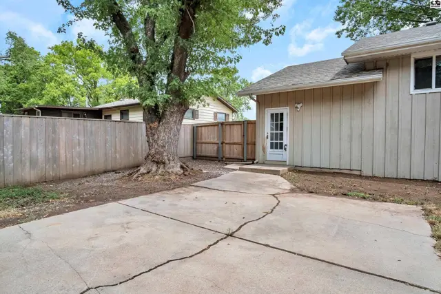Concrete patio leads to a backyard with a wooden fence and mature tree.
