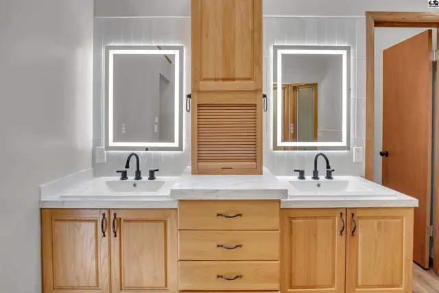 This bathroom features a double vanity with two illuminated mirrors and wood cabinetry.
